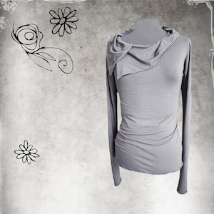 May include: A light gray long-sleeved top with an asymmetrical cowl neck design. The top is displayed on a mannequin and has a fitted silhouette. The background features floral sketches in black.