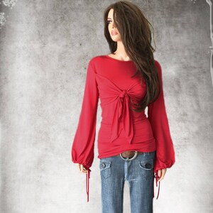 May include: A red long-sleeved shirt with a tie at the waist and a gathered detail at the cuffs. The shirt is worn with blue jeans and a brown belt.
