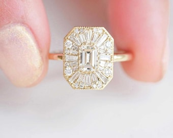 May include: A gold ring with a rectangular center stone and a halo of smaller, baguette-cut diamonds. The ring has a vintage-inspired design with intricate detailing around the setting. The band is thin and delicate.