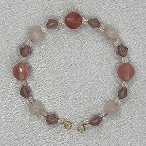 Peut inclure: Un bracelet de perles roses, violettes et transparentes. Le bracelet est composé de perles rondes et facettées enfilées sur un fil doré, avec une fermeture. Les perles sont dans des tons roses et violets. Circonférence d'environ 18 cm.