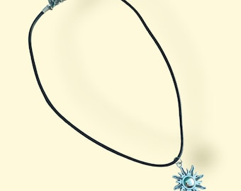 May include: A black cord necklace with a silver-colored sun pendant. The sun pendant has a detailed design with a central circular element. The necklace is displayed against a light yellow background.