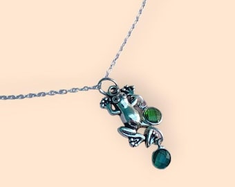 Könnte beinhalten: Halskette mit einem silberfarbenen Frosch-Anhänger. Der Anhänger hat detaillierte Merkmale und ist mit zwei grünen Edelsteinen verziert. Die Kette ist filigran und silberfarben. Der Anhänger vor einem sanften, pfirsichfarbenen Hintergrund.