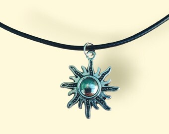 May include: A silver-toned sun pendant necklace with a black cord. The sunburst pendant has a central, reflective, circular element. The necklace is displayed against a light yellow background, creating a contrast with the dark cord and pendant.