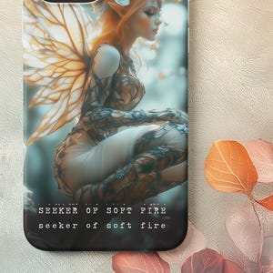 May include: A phone case featuring a fantasy art design of a fairy with orange hair, wings, and floral accents. The case has a black border and the text "Seeker of Soft Fire". The overall color scheme is soft and ethereal.