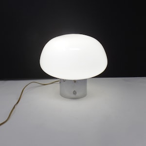 May include: A white mushroom-shaped table lamp with a rounded shade and a cylindrical base. The lamp is made of glass and metal, and has a power cord. The lamp is designed to provide soft, ambient lighting.