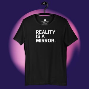 May include: Black crew neck t-shirt with the white text "REALITY IS A MIRROR." The shirt is hanging on a black hanger against a purple and pink gradient background.