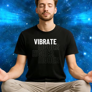 May include: Black t-shirt with the words "VIBRATE HIGHER HIGHER HIGHER" in white and outlined text. The person in the image is wearing the shirt and is sitting with their legs crossed and eyes closed. The background is a blue and white starburst pattern.