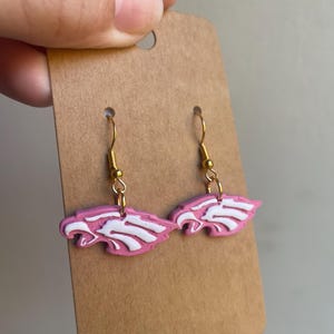 May include: A pair of pink and white earrings with a stylized eagle design. The earrings are held by a gold-colored hook and are attached to a brown cardboard display card. The earrings are a light pink color with white accents.