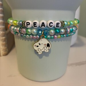 May include: Stacked beaded bracelets with the word "PEACE" spelled out in white letter beads. The bracelets feature a variety of colorful beads, including iridescent pearls and clear crystals. A charm of a cartoon character is attached to the bottom bracelet.