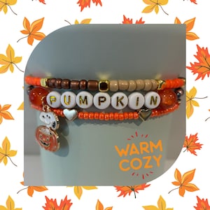 May include: A stack of three beaded bracelets featuring orange, brown, and beige seed beads, white letter beads spelling PUMPKIN, gold heart charms, and a metal charm of a character sitting in a pumpkin. The text WARM COZY is visible.