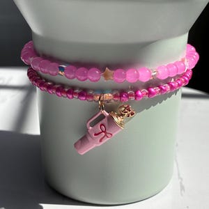 May include: Two pink beaded bracelets stacked on top of each other. The top bracelet has larger, round beads and a small star charm. The bottom bracelet has smaller beads and a pink charm shaped like a watering can with a pink ribbon.