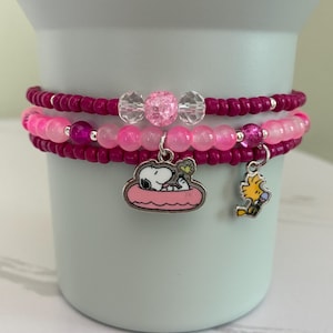 Op de afbeelding: Een set van drie gestapelde kralenarmbanden in roze en magentatinten. De armbanden zijn voorzien van kleine ronde kralen, heldere gefacetteerde accenten en twee metalen bedels met Snoopy in een roze zwemband en Woodstock met een ijsje.