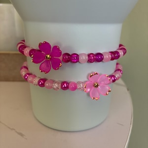 May include: Two pink beaded bracelets, each adorned with a large, vibrant flower charm. The bracelets feature a mix of pink beads in varying shades, creating a delicate and feminine accessory. The flower charms have gold accents.