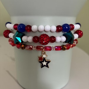 Op de afbeelding: Drie armbanden met rode, witte en blauwe kralen. De bovenste armband heeft witte, rode en blauwe kralen. De middelste armband heeft rode, witte en een groene stervormige kraal. De onderste armband heeft een sterbedel.
