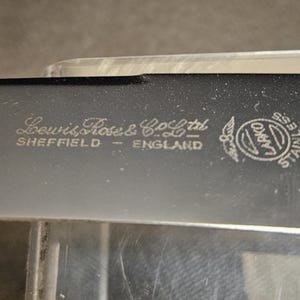 Pu&ograve; includere: Primo piano di una lama nera in acciaio inossidabile con la scritta "Lewis Rose & Co Ltd SHEFFIELD ENGLAND" e un logo circolare. La lama ha un bordo leggermente curvo e una finitura lucida.