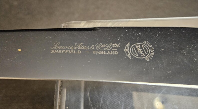 Pu&ograve; includere: Primo piano di una lama nera in acciaio inossidabile con la scritta "Lewis Rose & Co Ltd SHEFFIELD ENGLAND" e un logo circolare. La lama ha un bordo leggermente curvo e una finitura lucida.