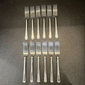 May include: A set of twelve silver forks arranged in two rows on a dark surface. Each fork has a long, slender handle with a decorative design near the base and four tines. The forks are made of a shiny, reflective metal.