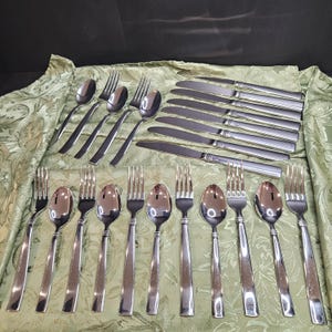 Può includere: Un set di posate in acciaio inossidabile, tra cui forchette, cucchiai e coltelli, disposti su una tovaglia verde fantasia. Le posate hanno un design semplice e moderno con una finitura lucida.