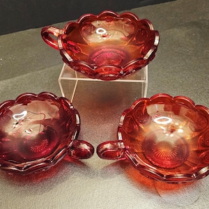 Könnte beinhalten: Drei antike rote Glasbecher mit Henkeln. Die Becher haben einen gewellten Rand und ein Sternmuster auf der Basis. Das Glas hat eine reflektierende, irisierende Qualität. Die Becher werden auf einem transparenten Acrylständer präsentiert.