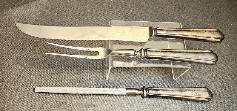 Pu&ograve; includere: Un set da intaglio in tre pezzi con manici argentati e lame in acciaio inossidabile. Il set comprende un coltello da intaglio, una forchetta da intaglio e un'acciarino. I manici hanno un design decorativo e scanalato.