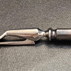 Pu&ograve; includere: Una forchetta da intaglio argentata con un'estremit&agrave; a due rebbi e un manico decorativo. La forchetta &egrave; tenuta da una mano e lo sfondo &egrave; una superficie scura e strutturata. La forchetta sembra essere vintage o antica.