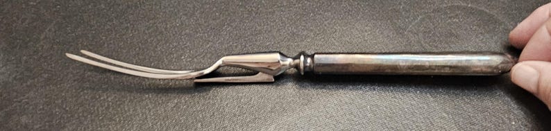 Pu&ograve; includere: Una forchetta da intaglio argentata con un'estremit&agrave; a due rebbi e un manico decorativo. La forchetta &egrave; tenuta da una mano e lo sfondo &egrave; una superficie scura e strutturata. La forchetta sembra essere vintage o antica.