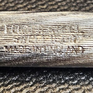 Pu&ograve; includere: Primo piano di un oggetto metallico argentato con il testo "FORGED STEEL STERLING" e "MADE IN ENGLAND" inciso sulla superficie. L'oggetto ha un aspetto strutturato, simile alle venature del legno.