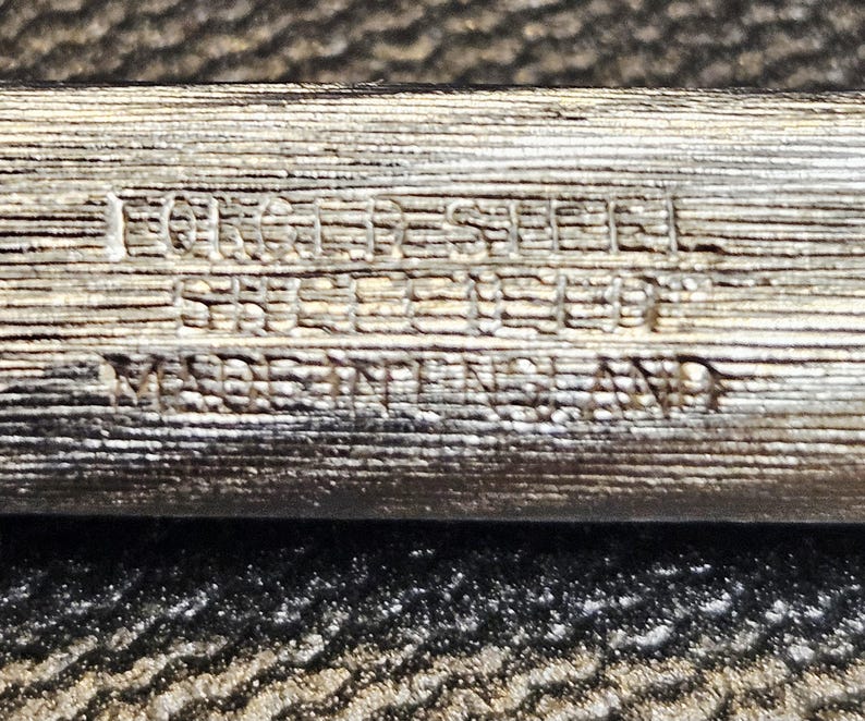 Pu&ograve; includere: Primo piano di un oggetto metallico argentato con il testo "FORGED STEEL STERLING" e "MADE IN ENGLAND" inciso sulla superficie. L'oggetto ha un aspetto strutturato, simile alle venature del legno.