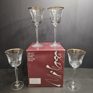 May include: Four clear crystal wine goblets with gold rims. The goblets are displayed with a red box that reads "MIKASA Full Lead Crystal 001 Goblet 4 Pieces". The goblets have a tall, slender stem and a wide bowl.