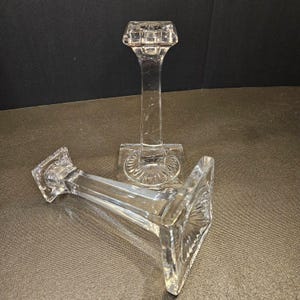 May include: Two clear glass candle holders. Each has a square base with a decorative pattern, a slender stem, and a square top. One is upright, the other is lying on its side. The candle holders are made of clear glass.