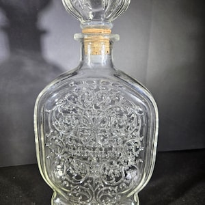 Puede incluir: Decantador de vidrio transparente con tapón decorativo. El decantador tiene un diseño ornamental en relieve y un tapón de corcho. El tapón está rematado con un elemento decorativo de vidrio acanalado. El decantador está vacío.