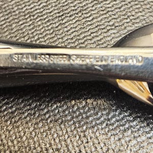 Pu&ograve; includere: Primo piano di un'utensile da portata lucido in acciaio inossidabile. Il testo "STAINLESS STEEL SHERRIFF ENGLAND" &egrave; inciso sul manico. L'utensile ha una superficie lucida e riflettente ed &egrave; posto su uno sfondo strutturato.
