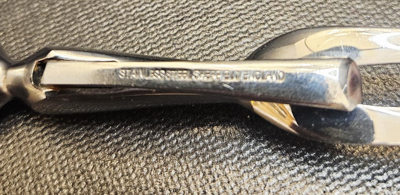 Pu&ograve; includere: Primo piano di un'utensile da portata lucido in acciaio inossidabile. Il testo "STAINLESS STEEL SHERRIFF ENGLAND" &egrave; inciso sul manico. L'utensile ha una superficie lucida e riflettente ed &egrave; posto su uno sfondo strutturato.