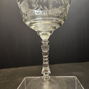 Puede incluir: Una copa de champán clara y vintage con diseños florales y de pájaros grabados. La copa presenta un cuenco ancho, un tallo facetado y una base circular. Los detalles grabados incluyen hojas y un pájaro.