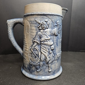 May include: A blue and white ceramic beer stein with a detailed relief of a man in historical attire. The stein features a handle and a light-colored rim. The design includes a man holding a barrel and a mug, set against a brick wall background.