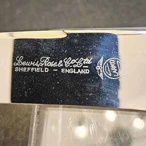 Pu&ograve; includere: Primo piano di un oggetto in metallo argentato con la scritta bianca "Lewis Rose & Co Ltd SHEFFIELD - ENGLAND". &Egrave; visibile anche un logo circolare con la parola "LARKO". L'oggetto ha una superficie riflettente.