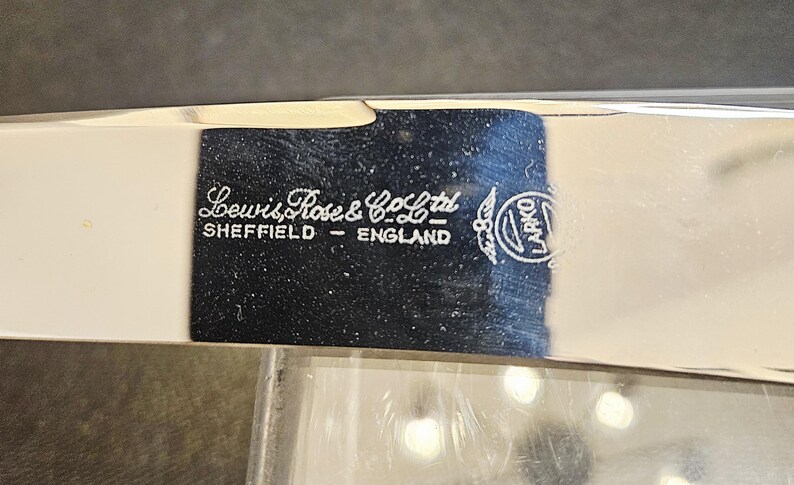 Pu&ograve; includere: Primo piano di un oggetto in metallo argentato con la scritta bianca "Lewis Rose & Co Ltd SHEFFIELD - ENGLAND". &Egrave; visibile anche un logo circolare con la parola "LARKO". L'oggetto ha una superficie riflettente.