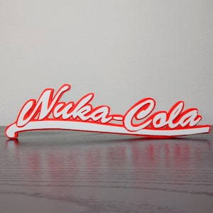 May include: A red and white Nuka-Cola logo, a stylized sign from the Fallout video game series. The sign is in a cursive font with a red outline and white fill, resting on a dark wooden surface.