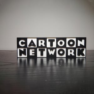 May include: A black and white sign that reads "CARTOON NETWORK". The letters are white and set against a black background, with the sign resting on a dark surface. The sign is a decorative item.