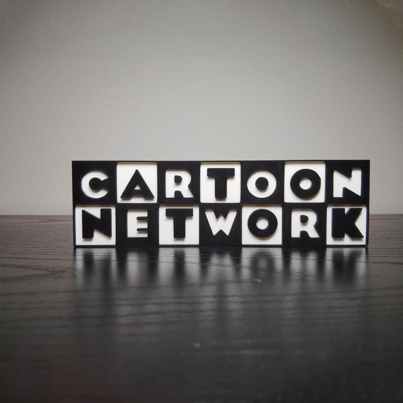 May include: A black and white sign that reads "CARTOON NETWORK". The letters are white and set against a black background, with the sign resting on a dark surface. The sign is a decorative item.