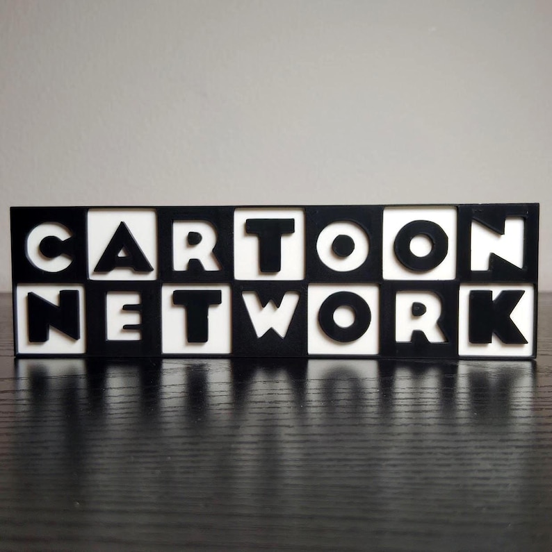 May include: A black and white sign that spells out "CARTOON NETWORK". The letters are black and set against a white background. The sign is rectangular and sits on a dark surface.