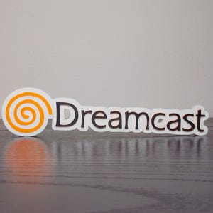 May include: A white sticker with the word "Dreamcast" in black letters. The "Dreamcast" text is next to a spiral design in orange and white. The sticker is on a dark wooden surface.