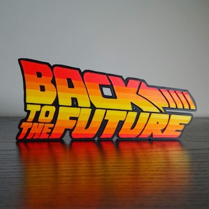 May include: A colorful Back to the Future logo in red, orange, and yellow, with a black outline. The text reads "BACK TO THE FUTURE" in a stylized font, with a design resembling a flux capacitor.