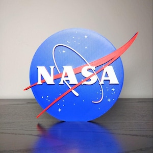 May include: A blue, white, and red NASA logo. The logo features a blue circle with white stars and the word "NASA" in white block letters. A red swoosh curves across the circle. The logo is displayed on a dark surface.