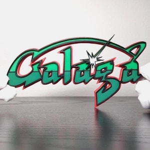 May include: A green and red layered sign with the word "Galaga" in a stylized font. The sign has a green outline with red accents and a starburst graphic. The sign is on a dark surface.