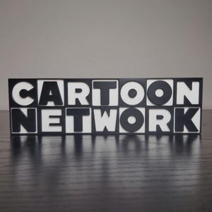 May include: A black and white sign that reads "CARTOON NETWORK". The letters are black, and the background is white. The sign is sitting on a dark surface.