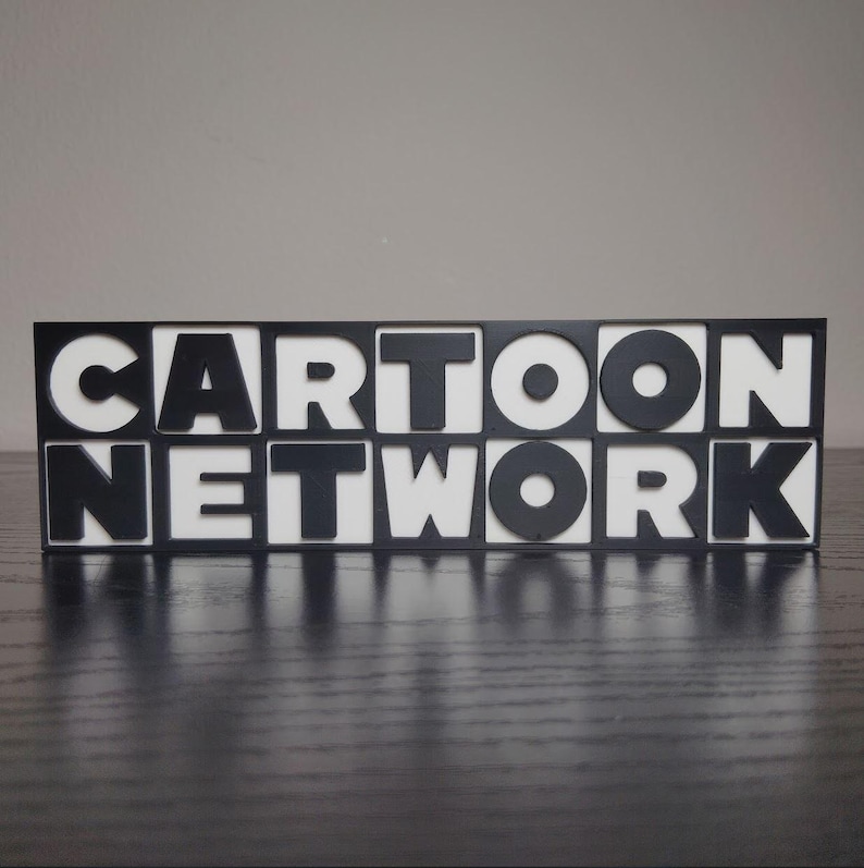 May include: A black and white sign that reads "CARTOON NETWORK". The letters are black, and the background is white. The sign is sitting on a dark surface.
