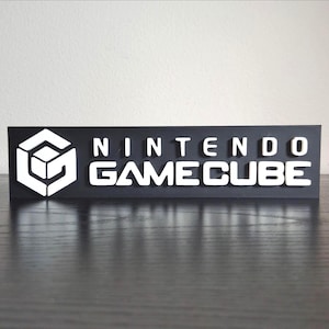 May include: A black rectangular sign with the Nintendo GameCube logo and text in white. The logo is a white cube within a cube. The text reads "NINTENDO GAME CUBE".