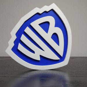 May include: A white and blue Warner Bros. logo, featuring the letters "WB" in a stylized design. The logo is set against a blue background and framed by a white border with a mountain-like top edge. The logo is on a dark surface.