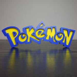 May include: A blue and yellow Pokemon logo, with the word "Pokemon" in a stylized font. The letters are yellow with a blue outline, and the logo is set against a dark background.